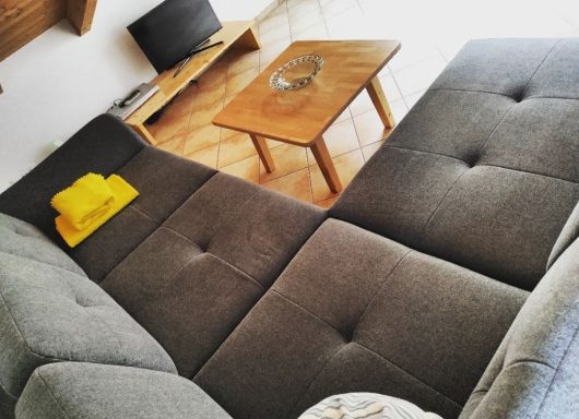 Wohnzimmer, graue Couch mit Kissen und Decken, ein Holztischchen und ein TV mit DVD Player.